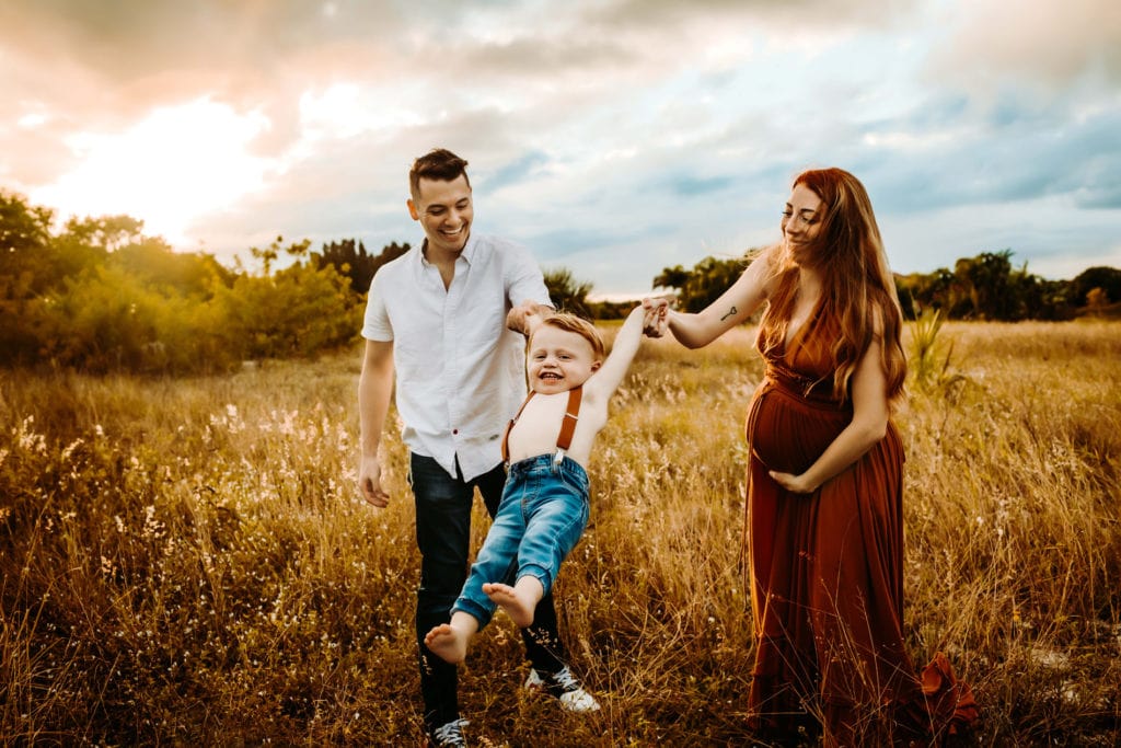 Family Photographer, mom and dad swing young son by his arms in a field
