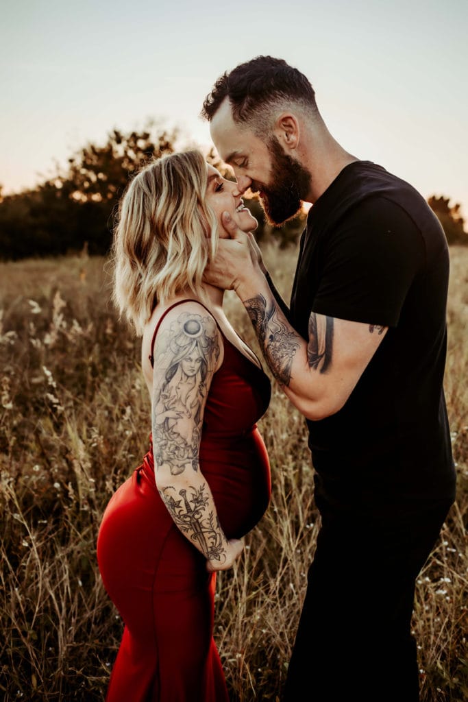 Maternity Photographer, husband draws close to expecting wife , they are standing in dry grassy field